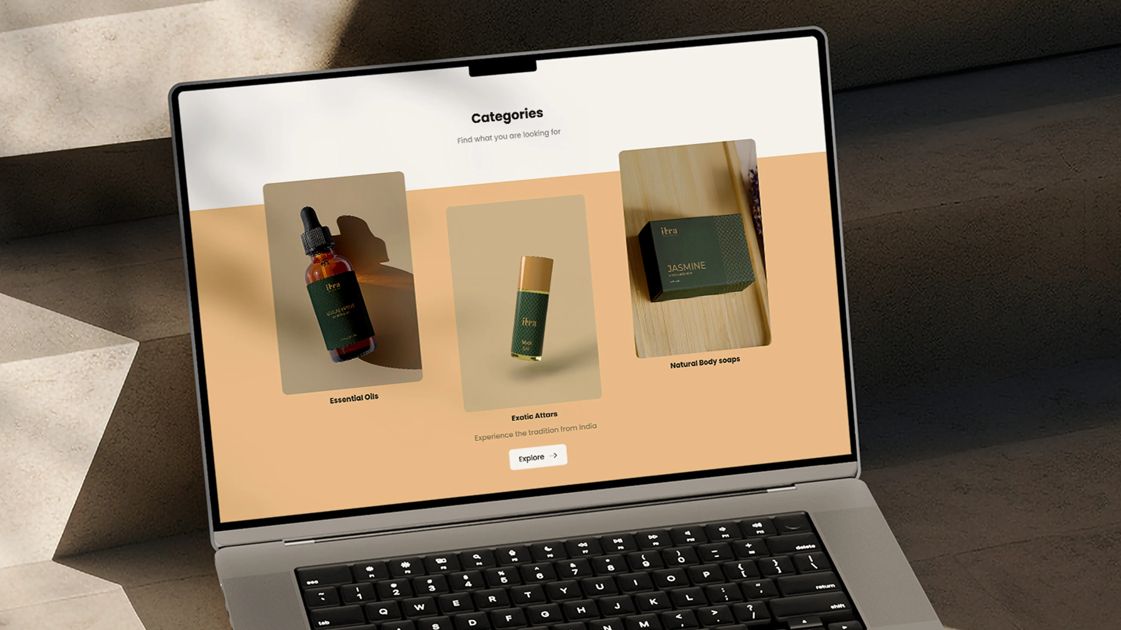 Itra website categories section on a MacBook screen showing three staggered cards for Essential Oils, Exotic Attars, and Natural Body Soaps on a warm sand background